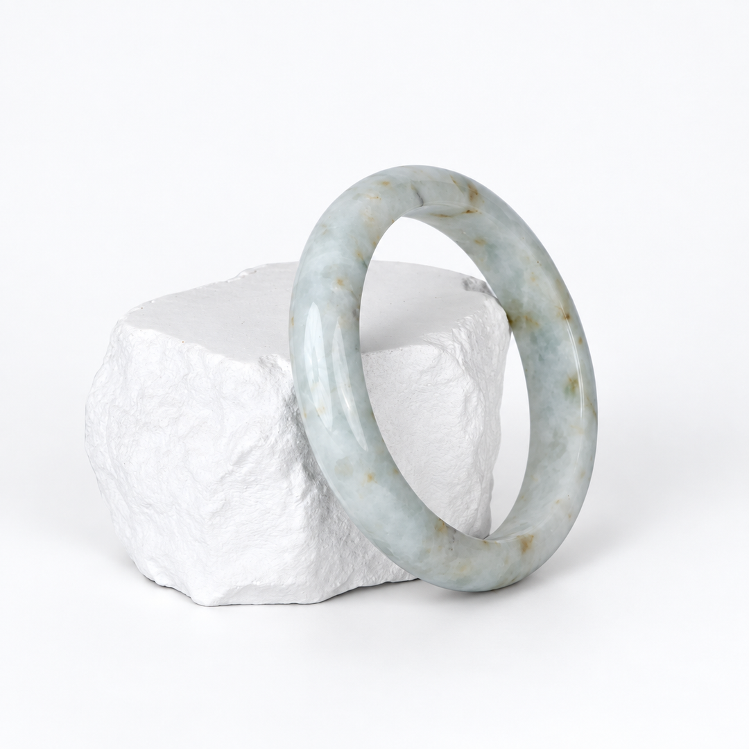 White jadeite bangle with small brown and grey spots on a white textured display rock, three-quarter view